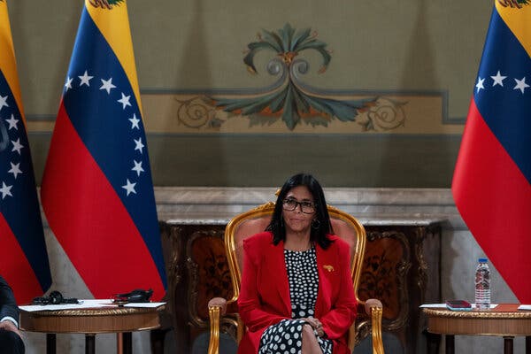 Delcy Rodríguez, la líder interina de Venezuela, está sentada en una silla dorada, flanqueada por dos banderas amarillas, azules y rojas. En primer plano hay unas mesitas con una botella de agua y un micrófono.