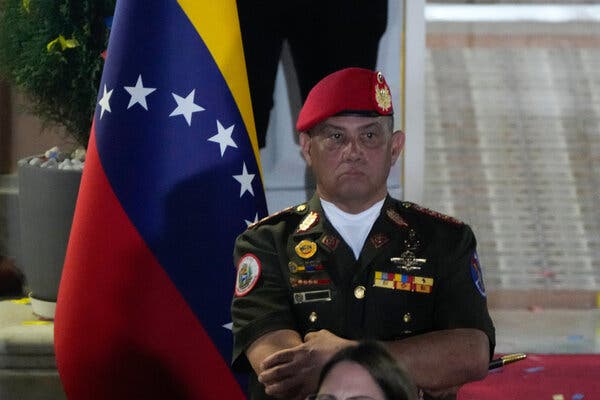 Una persona con uniforme militar y una boina roja, está de pie con aire severo delante de la bandera venezolana.